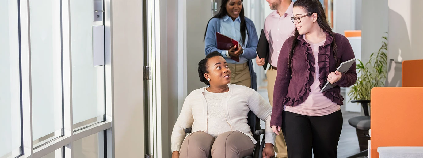 A women in a wheelchair and her female colleague talking as they move through a tidy hallway.
