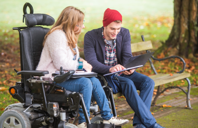 A young woman in a wheelchair and a man are in the park looking at a tablet and talking.
